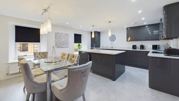 A modern kitchen with a central island, pendant lighting, and a dining area with a glass table and cushioned chairs. - Charming 3-Bedroom Home in the Heart of Stanhope (Stanhope)