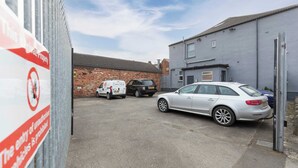 A parking area with a white car and a van, a brick building, and a grey building with a garage door. - Middlesborough City Centre Studio (Middlesbrough)