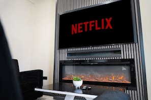 Ein modernes Wohnzimmer mit Kamin, einem Fernseher, der 'NETFLIX' zeigt, und einer kleinen Pflanze auf dem Kaminvorsatz.
