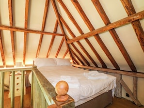 Quarto com teto inclinado, vigas de madeira, cama com roupa de cama branca e uma mesa de cabeceira de madeira com gavetas.