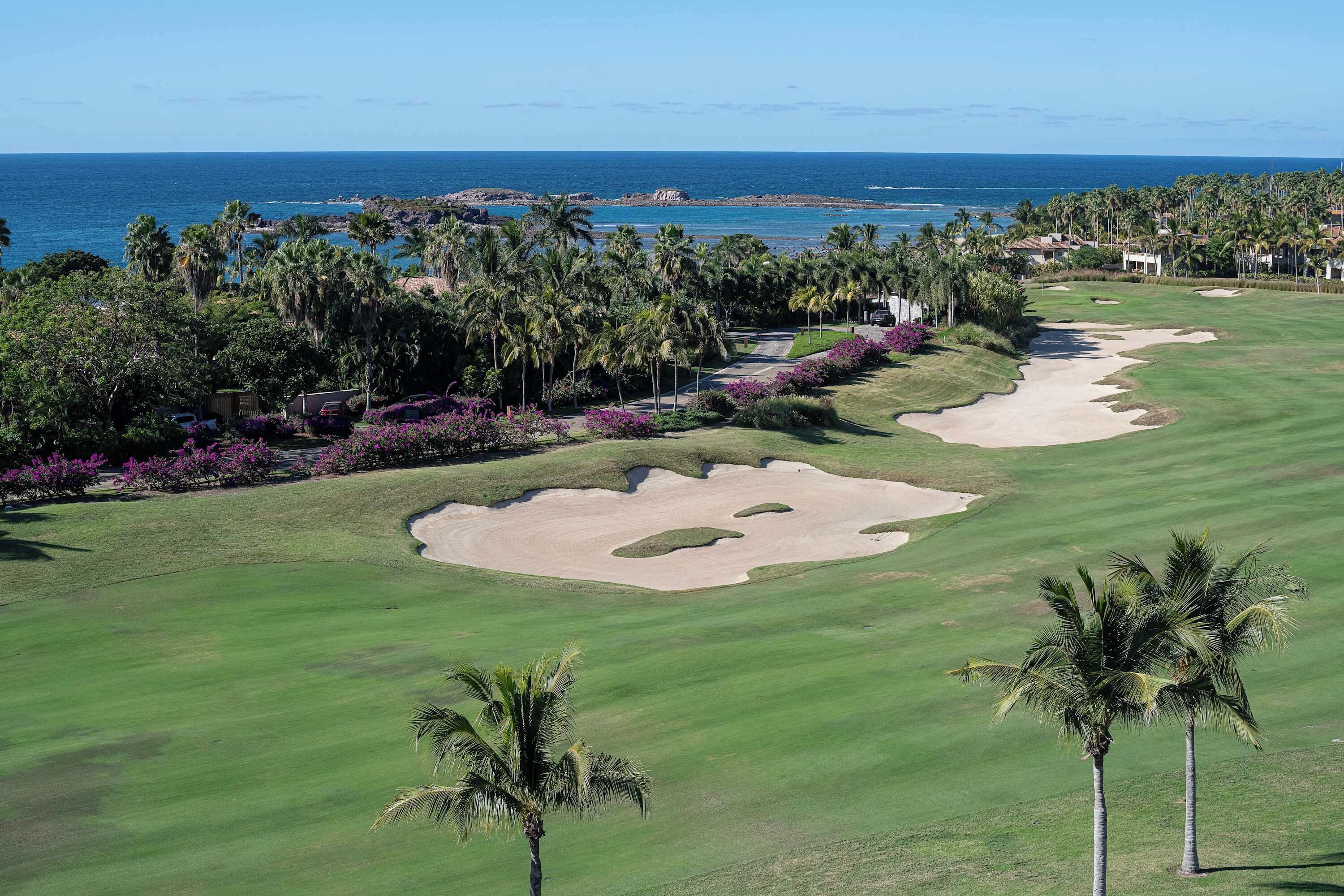 A golf course with sand bunkers, palm trees, and a view of the ocean.