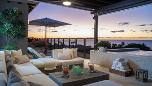 A modern outdoor seating area with a view of the ocean at sunset, a lounge chair, a sofa, and a coffee table with decorative items. - PH Azure: 4BR Oceanview Penthouse w/ Private Heated Infinity Pool & Rooftop (Punta Mita)