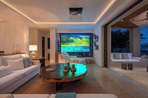 Ein modernes Wohnzimmer mit großem Flachbildfernseher, weißen Sofas, einem Couchtisch und Blick auf eine tropische Landschaft.