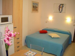 Ein Schlafzimmer mit einem Bett, einem Sessel, einem Tisch, einer Mikrowelle und einer Vase mit Blumen.