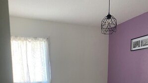 A single bed in a room with a window, a light fixture, and a wall with a picture. - Charming and cosy T2 near the beach, parking space (Sète)