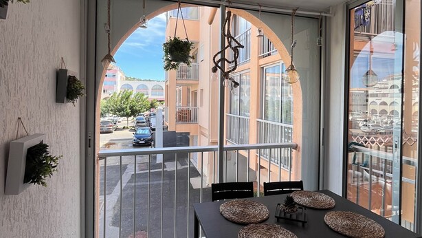 A balcony with a table and chairs, a hanging plant, and a view of buildings and parked cars. - Charming and cosy T2 near the beach, parking space (Sète)