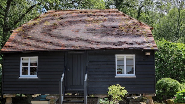 A black wooden house with a tiled roof, two windows, a door, and a small porch area with steps leading to the entrance. - The Granary (Petworth)