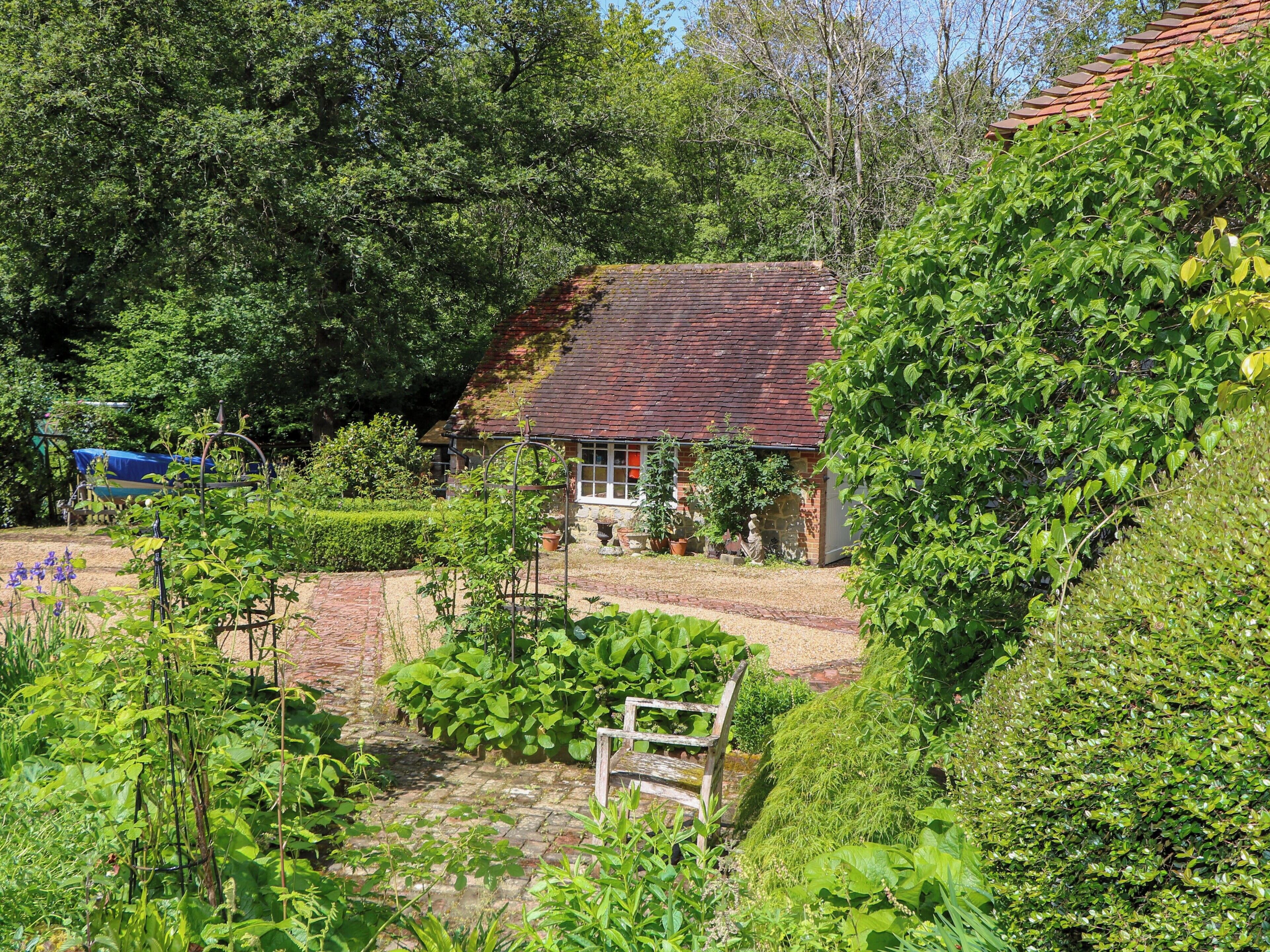 Una casa rústica rodeada de abundante vegetación, un jardín con un banco de madera y un cielo azul despejado.