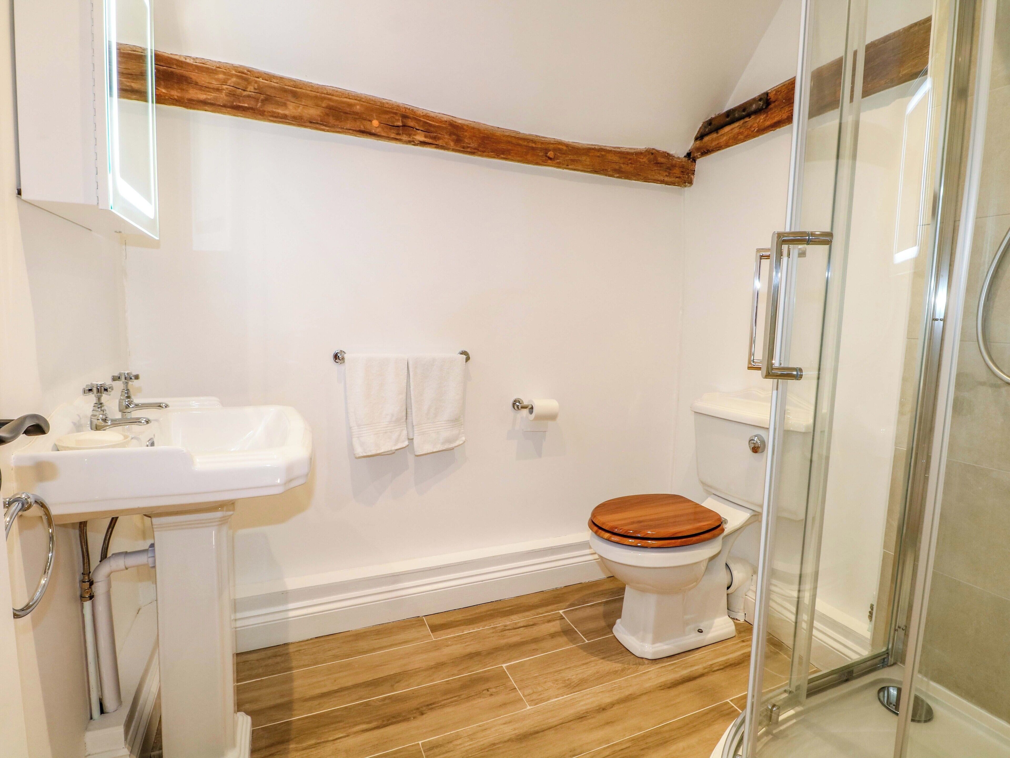 A bathroom with a wooden floor, white walls, a white sink, a toilet, and a glass shower enclosure.