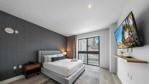 Moderni hotellihuone, jossa on suuri sänky, seinälle asennettu televisio, yöpöytä ja näkymä kaupunkiin ikkunasta.