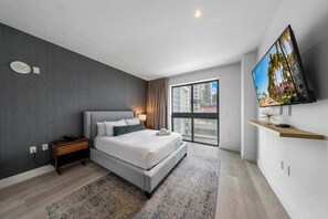 Ein modernes Hotelzimmer mit einem großen Bett, einem an der Wand befestigten Fernseher, einem Nachttisch und Blick auf die Stadt durch das Fenster.