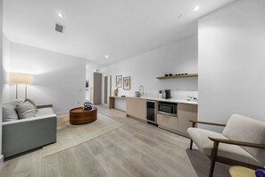 Ein modernes Wohnzimmer mit einer Couch, einem Couchtisch und einem Sessel. Es gibt eine Küchenzeile mit Mikrowelle, Backofen und Spüle.