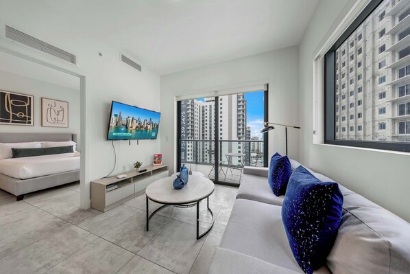 A modern living room with a sofa, a coffee table, a TV, and a balcony with a city view. - Luxe 1BR Balcony • Near FIFA Fan Fest (Miami)