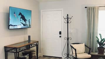 Una sala de estar con un televisor montado en la pared, una puerta blanca, un perchero, un sillón, una mesita y una planta en maceta.