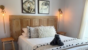 Ein Schlafzimmer mit einem Holzkopfteil, weißer Bettdecke, zwei Wandlampen und zwei gerahmten Kunstwerken über dem Bett.