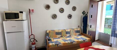 A compact room with a bed, a microwave, a refrigerator, a fire extinguisher, and a small table with a tablecloth.