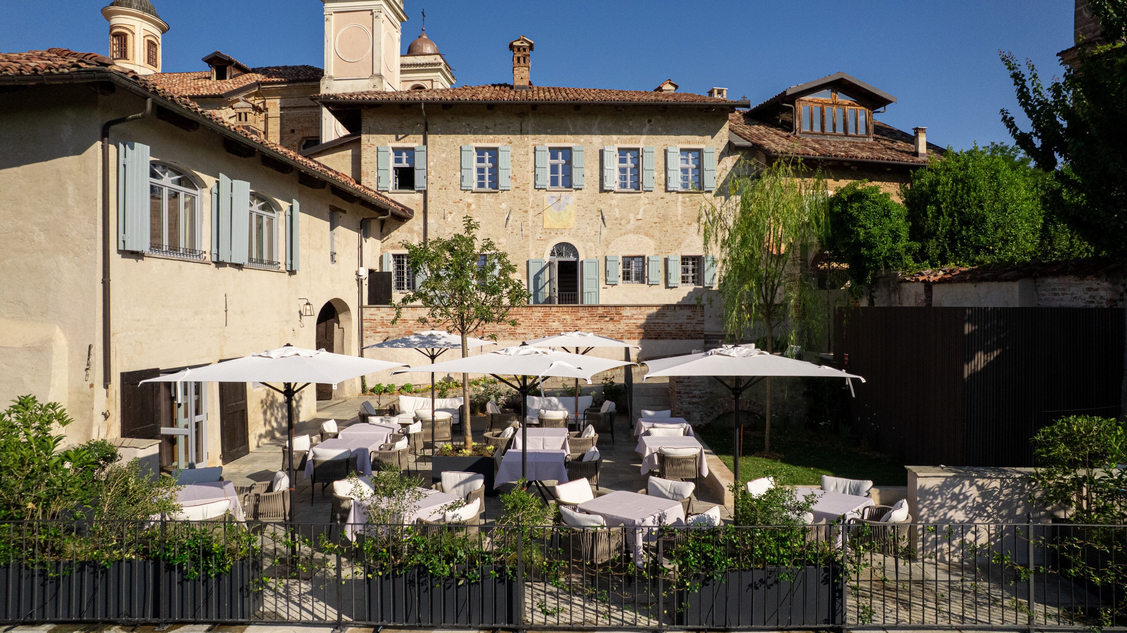 Une terrasse extérieure avec des tables et des chaises, entourée d’un mur de pierre et bordée d’un bâtiment doté de nombreuses fenêtres et d’un clocher.