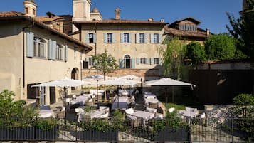 Une terrasse extérieure avec des tables et des chaises, entourée d’un mur de pierre et bordée d’un bâtiment doté de nombreuses fenêtres et d’un clocher.