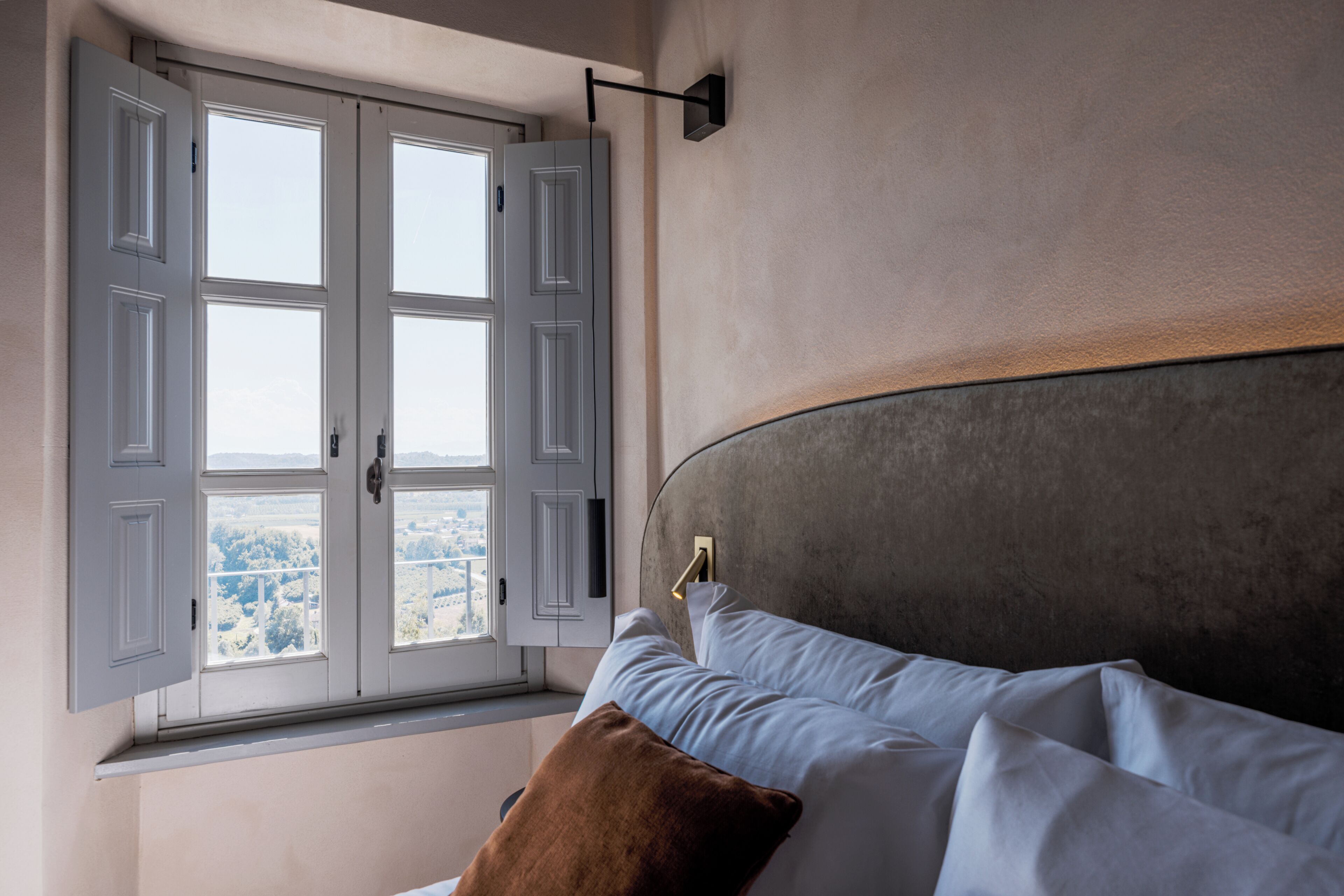 Quarto com uma janela grande, cama com roupa de cama branca e travesseiro marrom, e luminária fixada na parede.