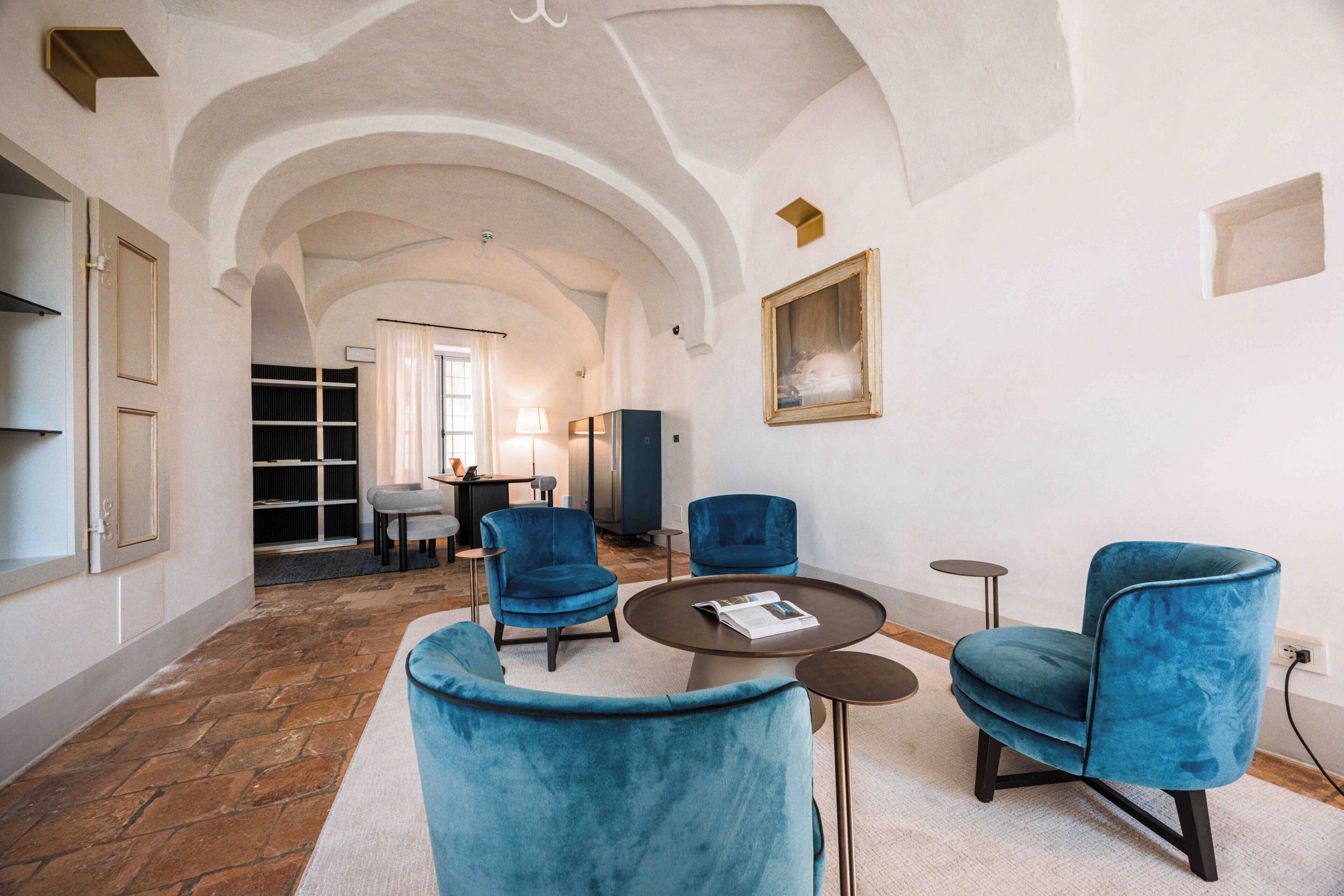 A hotel lobby with a stone floor, blue armchairs, a round table, and a painting on the wall.