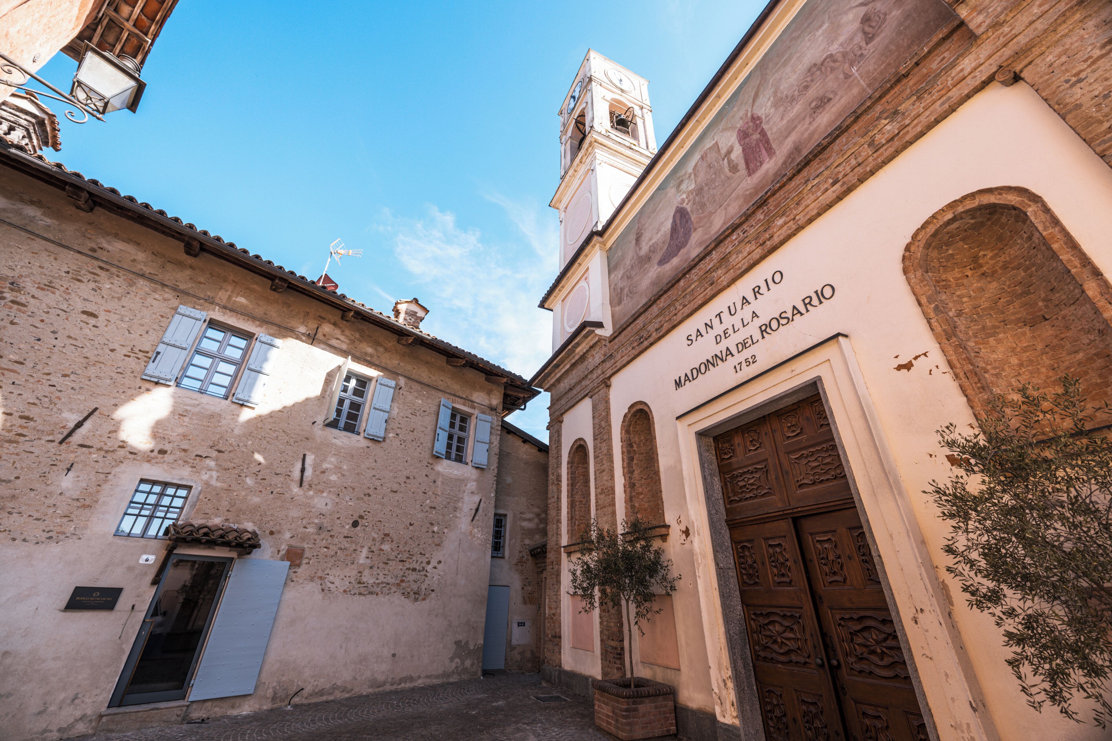 A narrow street with buildings featuring traditional architecture, including a bell tower and a building with a sign reading 'SANTUARIO DELLA MADONNA DEL ROSARIO 1752'.