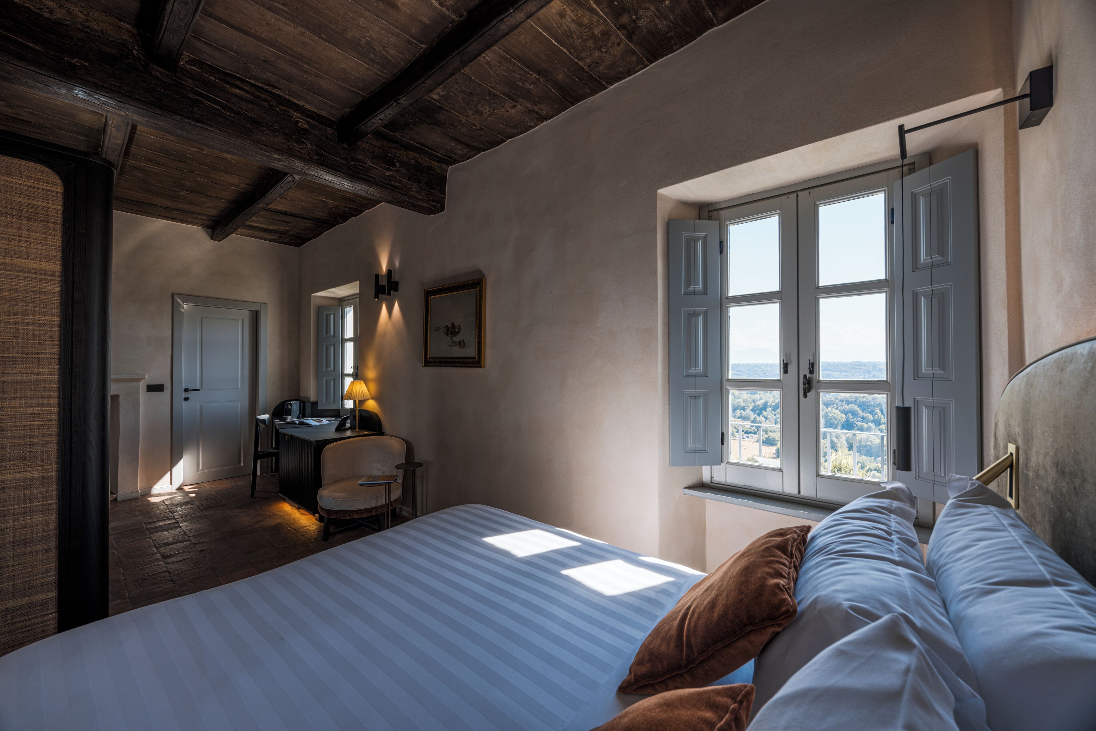 Een slaapkamer met een groot bed, een bureau en een stoel. Er is een raam met luiken en uitzicht op de omgeving.
