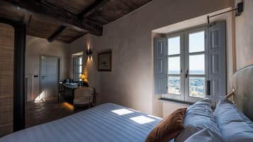 Een slaapkamer met een groot bed, een bureau en een stoel. Er is een raam met luiken en uitzicht op de omgeving.