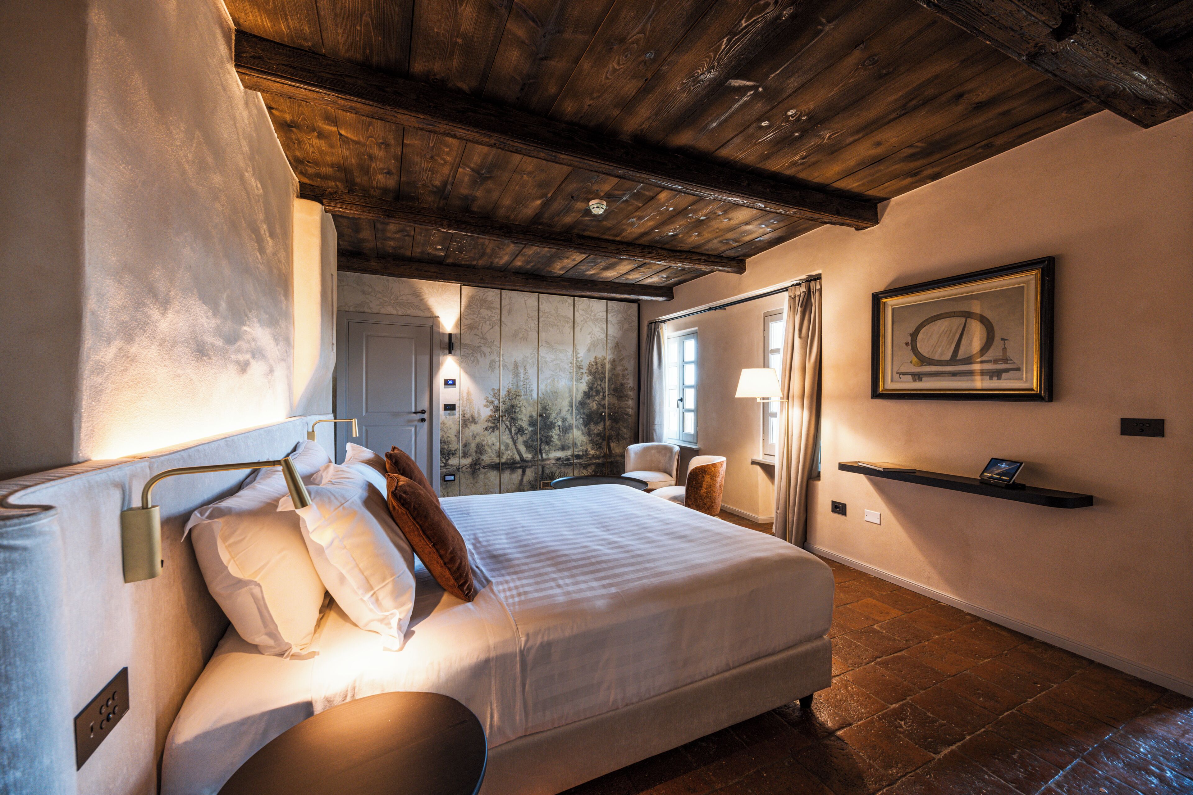 A bedroom with a large bed, a wooden ceiling, a painting on the wall, and a bedside table with a lamp.