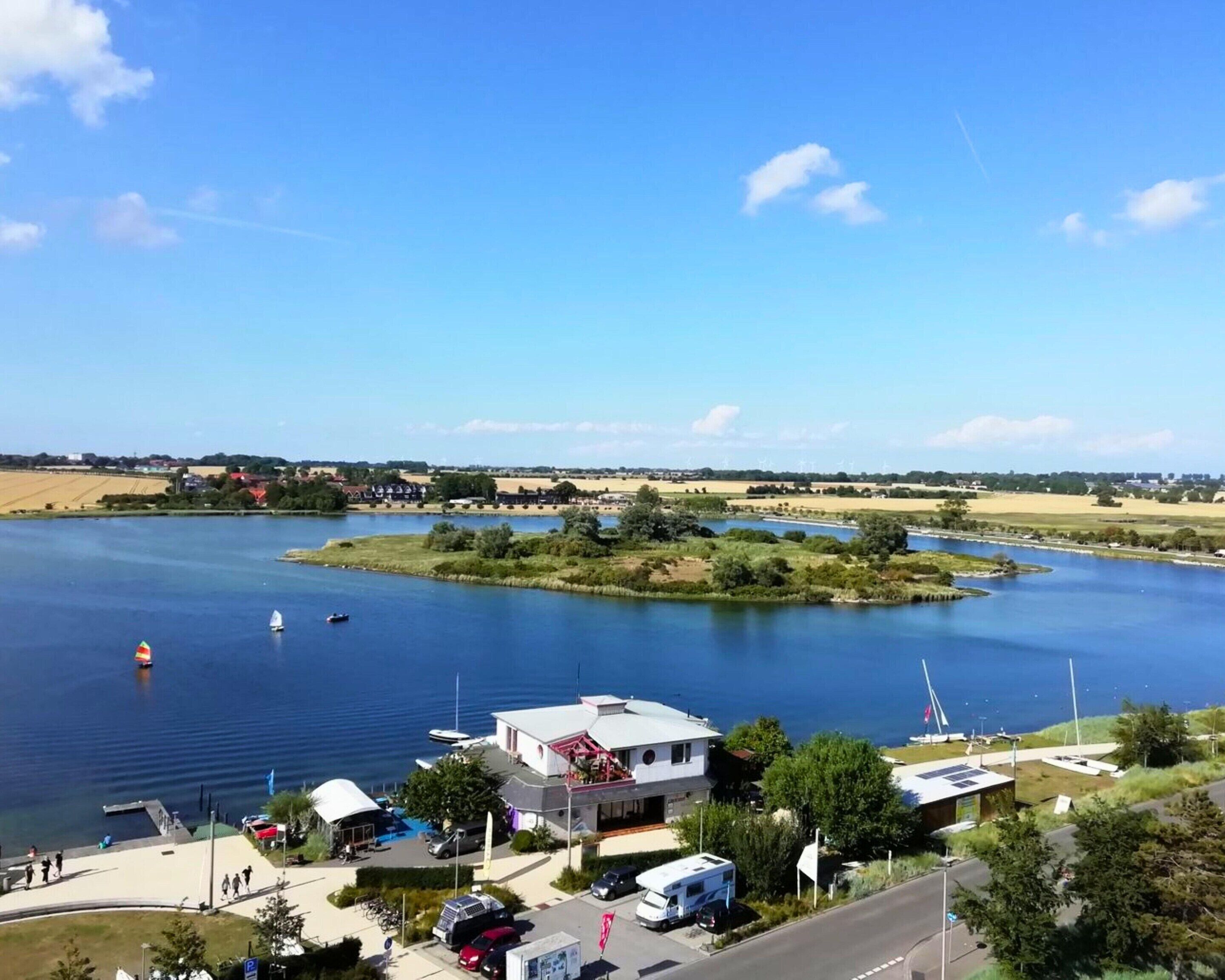 A lakeside property with a swimming pool, a parking area, and a view of the lake and distant wind turbines.