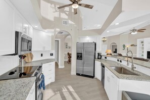 A modern kitchen with white cabinetry, stainless steel appliances, granite countertops, and a ceiling fan. - VRCC Villa Casa del Sol (Cape Coral)