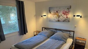 Een tweepersoonsbed met grijze beddengoed, een klein nachtkastje, een wandlamp en een raam met gordijnen.