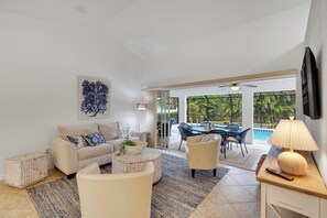 Ein Wohnzimmer mit einer Couch, einem Couchtisch und einem Essbereich mit Stühlen und einem Tisch. An der Wand ist ein Fernseher angebracht und es gibt einen Deckenventilator.