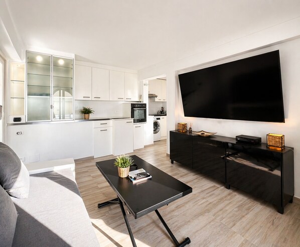 A modern kitchen with white cabinets, a large flat-screen TV, and a dining area with a black table and grey sofa.