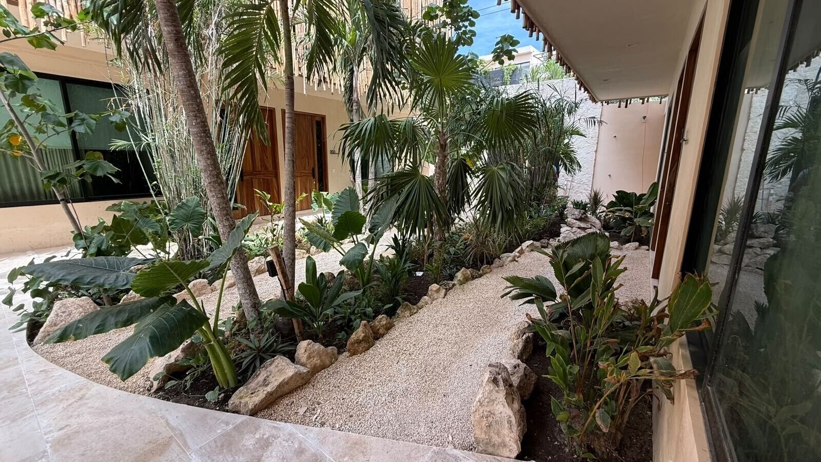 Un'area esterna curata con aiuole, un vialetto sterrato e diverse piante tropicali, con un edificio dalle ampie finestre.