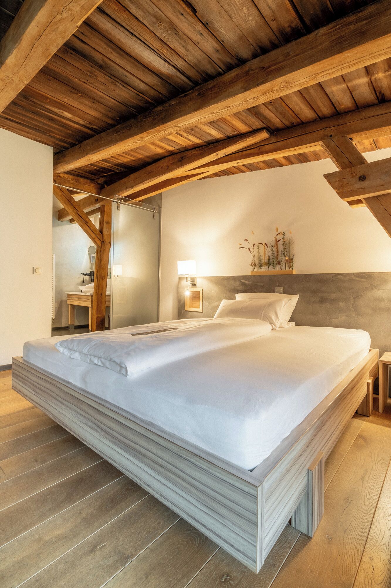 Une chambre avec un grand lit, des poutres en bois, une table de chevet et une lampe fixée au mur.