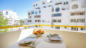 A balcony with a table set for two, a bowl of fruit, and a view of white buildings.