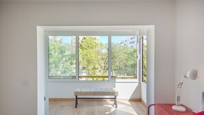 A room with a large window, a bench, a desk with a lamp, and a bed.