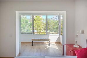 Ein Zimmer mit großem Fenster, einer Sitzbank, einem Schreibtisch mit Lampe und einem Bett.