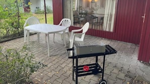 An outdoor patio with a table and chairs, a grill cart, and a view of the interior through large windows. - Vacation village Seddiner See - Bungalow 2 (Seddiner See)