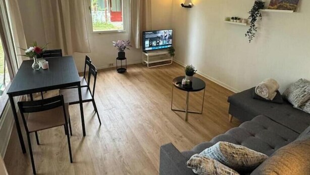 A living room with a sofa, a TV, a dining table with chairs, a small round table with a plant, and a window with curtains. - Vacation village Seddiner See - Bungalow 2 (Seddiner See)