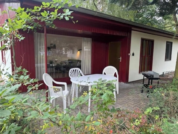 A covered outdoor seating area with a table and chairs, a glass door entrance, and a small table with items on it.