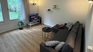 A living room with a sofa, a TV on a stand, a small table, and a window with curtains.