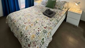 Quarto com colcha florida, travesseiro verde, pintura e mesa de cabeceira com abajur.