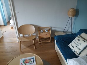 Et værelse med trægulv, seng med blåt sengetøj, et flettet natbord, en træstol og en gulvlampe.
