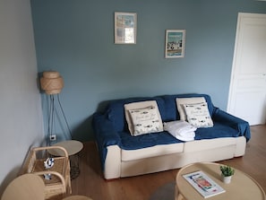 Une pièce avec un canapé bleu, deux coussins ornés de motifs, une petite table sur laquelle repose un magazine, une chaise en osier et une lampe fixée au mur.