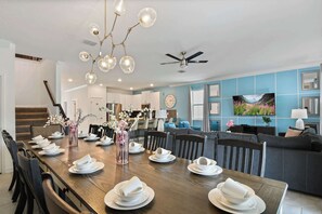 Ein Esszimmer mit einem langen Tisch, gedeckt für eine Mahlzeit, einem Deckenventilator und einer Küche im Hintergrund.