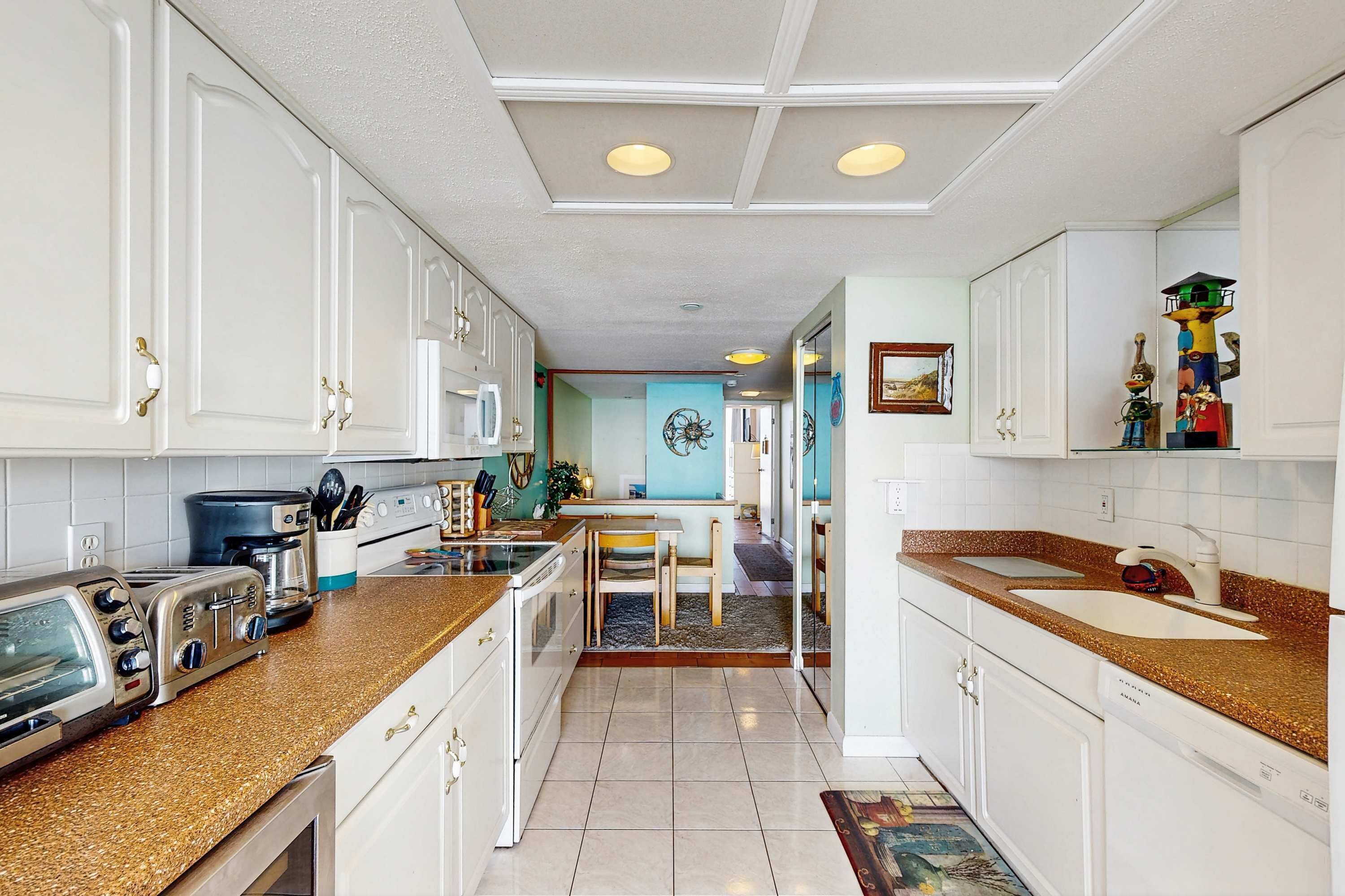 Uma cozinha com armários brancos, bancada de granito, espaço de jantar com mesa e cadeiras, e corredor com parede azul e espelho decorativo.