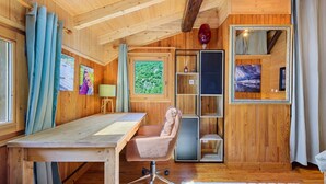 Interior de una cabaña de madera con un escritorio, una silla de oficina, estanterías y un ventanal grande.