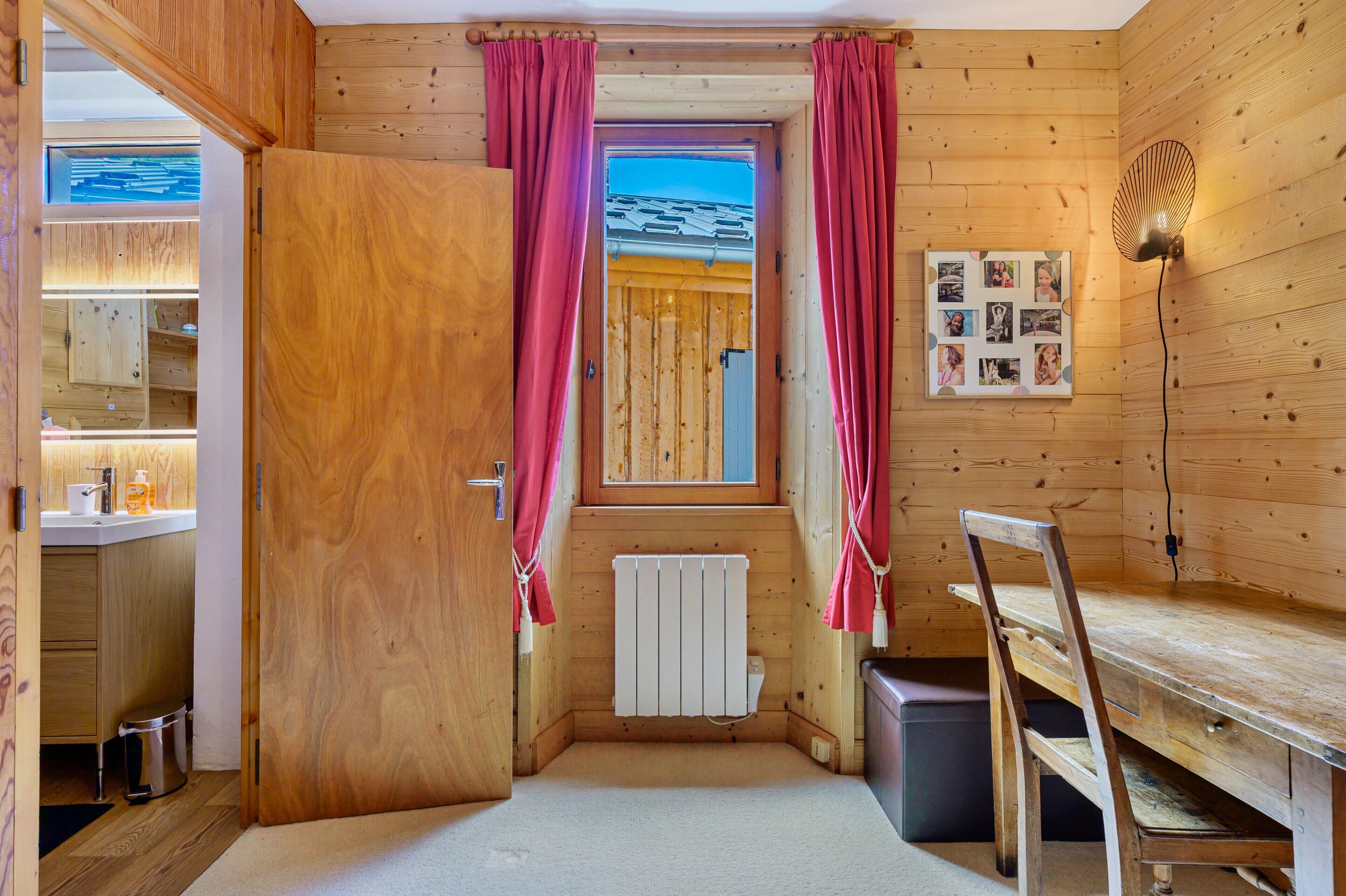 Ein Holzblockhaus-Interieur mit Holztür, Fenster mit Vorhängen, Heizkörper, Schreibtisch und einer Wandleuchte.