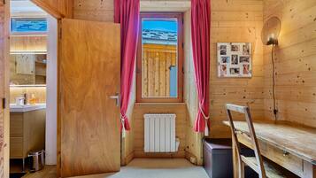 Ein Holzblockhaus-Interieur mit Holztür, Fenster mit Vorhängen, Heizkörper, Schreibtisch und einer Wandleuchte.
